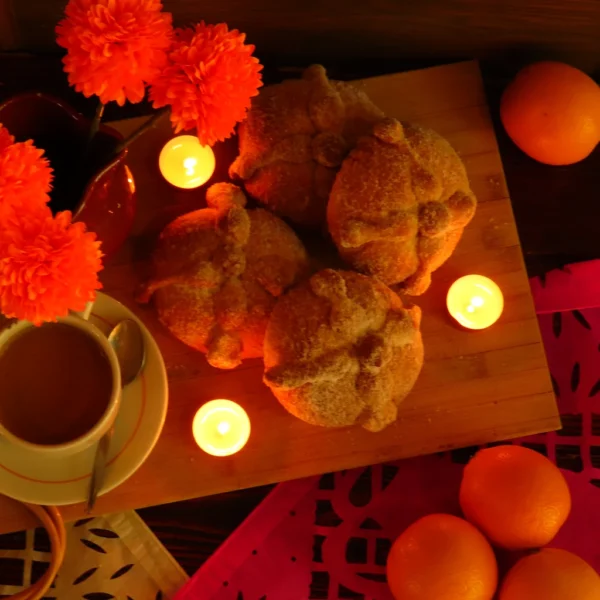 Cuatro panes de muerto sobre una tabla de madera, junto a flores naranjas, velas y una taza.