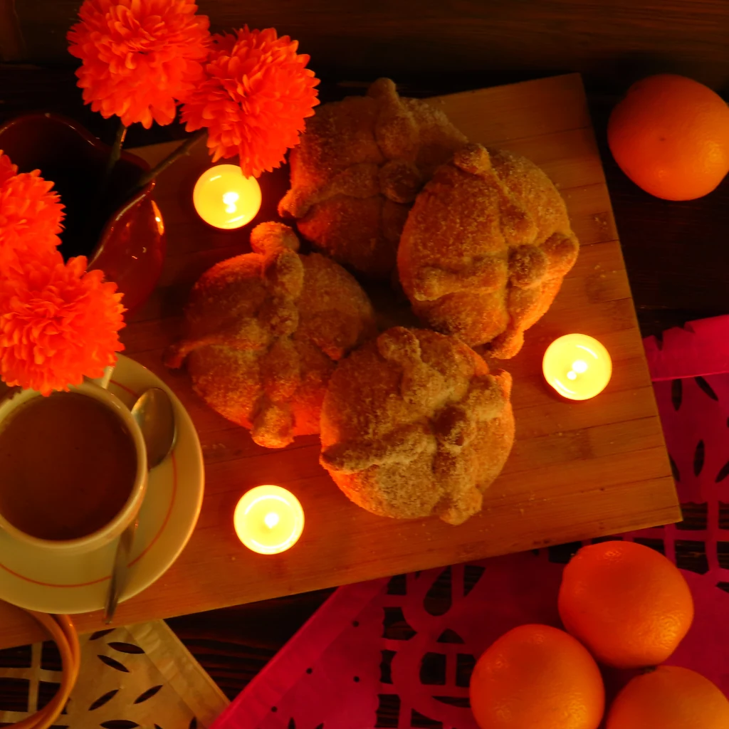 Cuatro panes de muerto sobre una tabla de madera, junto a flores naranjas, velas y una taza.
